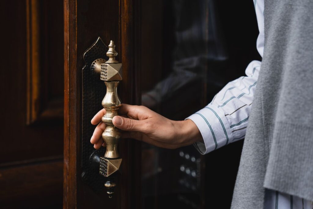Close-up of an ornate brass handleset on a dark wood door, a hand grasping the elegant hardware
