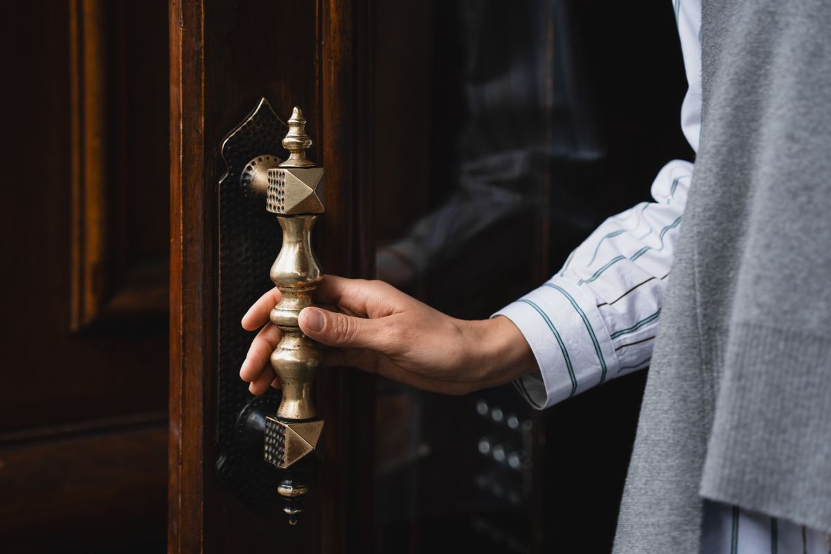 Close-up of an ornate brass handleset on a dark wood door, a hand grasping the elegant hardware