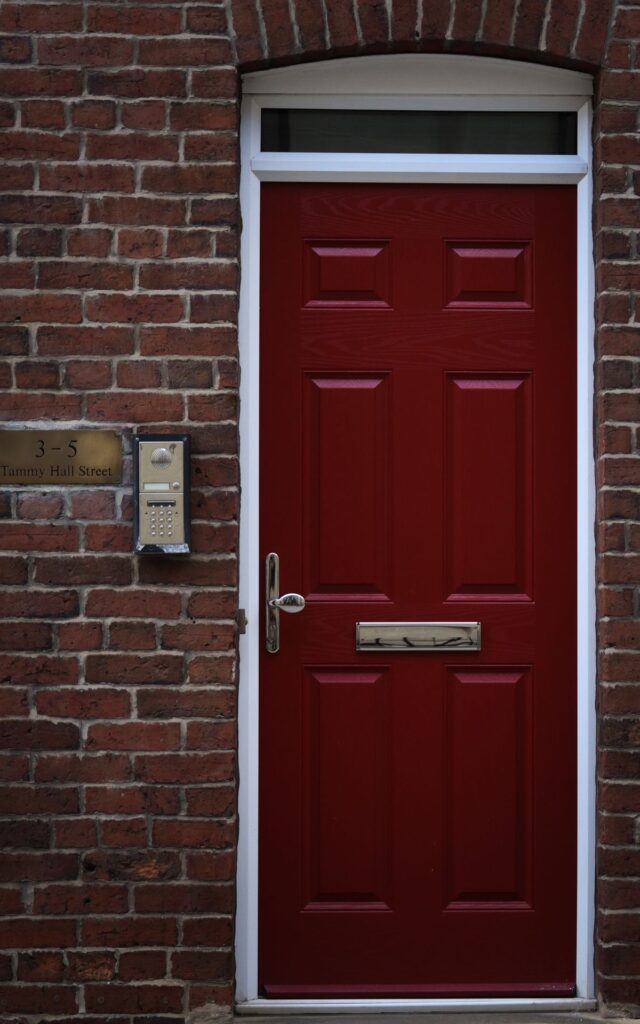 Intercom panel with keypad beside a red building entry door on a brick facade